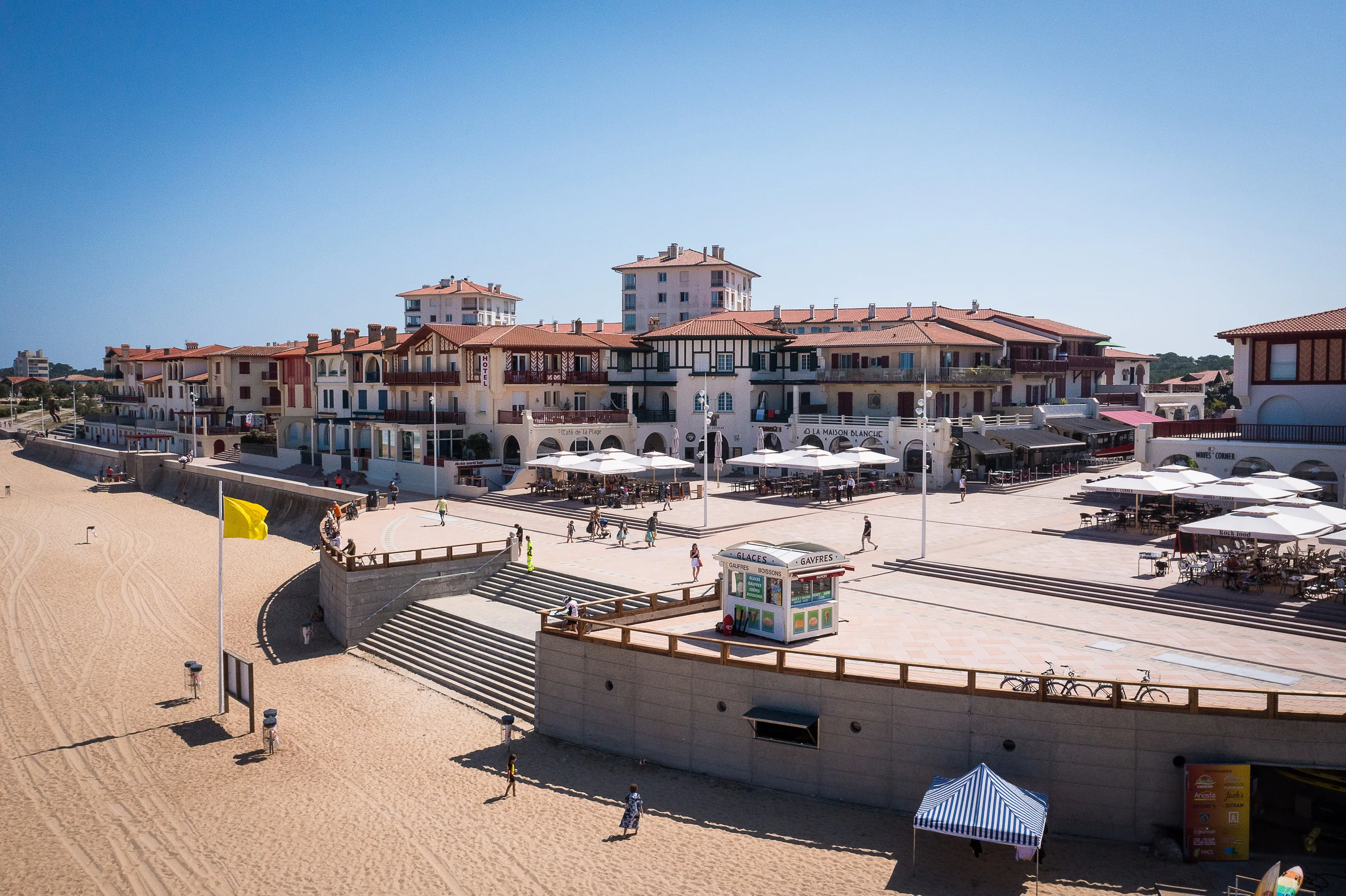 Vue aérienne panoramique de la plage des Landais à Hossegor prise par drone sous un ciel bleu dégagé, montrant le sable doré bordé de dunes couvertes de végétation, les vagues de l’océan Atlantique déferlant sur le rivage, des surfeurs présents dans l’eau, et en arrière-plan le village d’Hossegor niché au cœur des Landes, baigné par la lumière du soleil et offrant une atmosphère estivale et dynamique propre à la côte landaise.